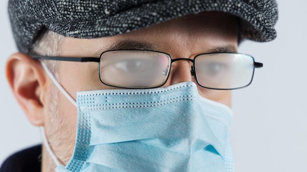 A man with a hat and glasses, wearing a surgical mask, has fogged glasses due to breathing while masked.